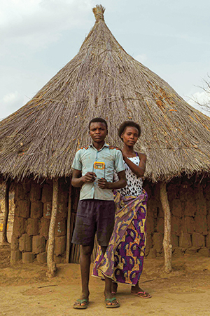 Derrick and his mother standing in front of a hut, holding up a solar lamp