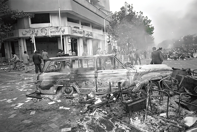 Black & white photo of the burned remains of two vehicles in a city street