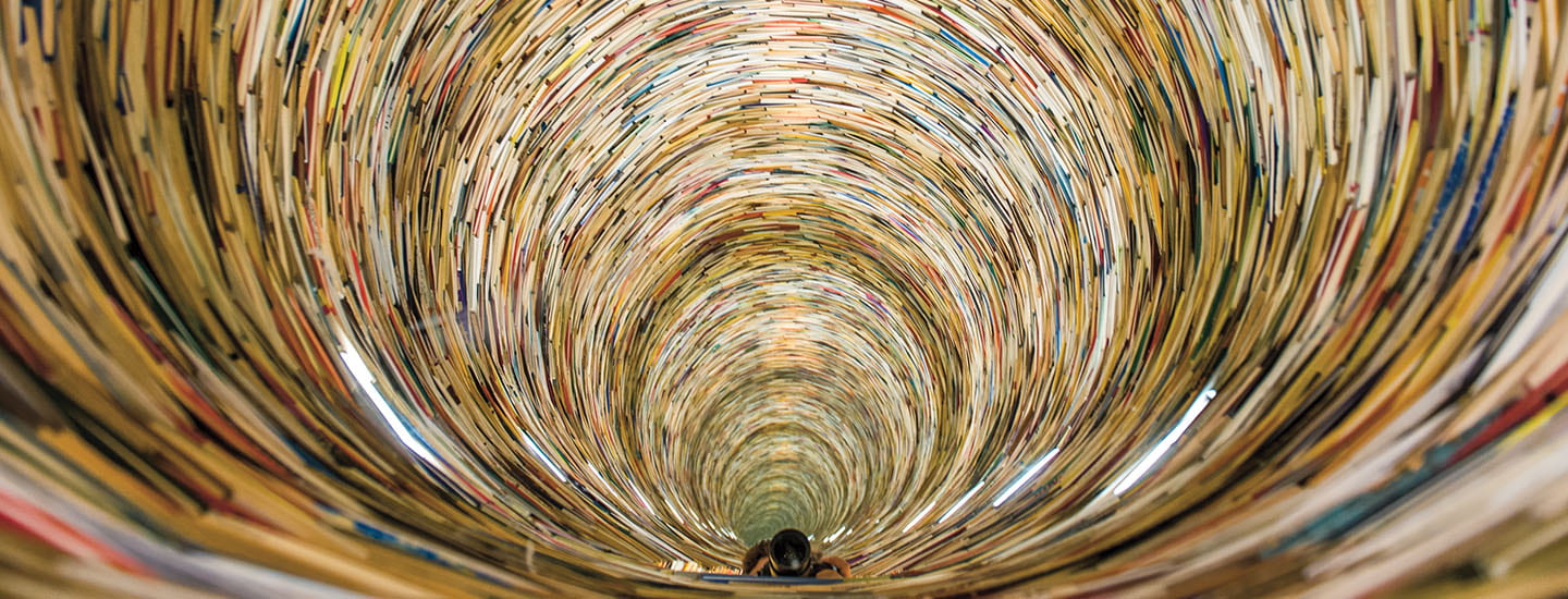 A person stands at the base of a towering cylindrical tunnel lined with stacked books