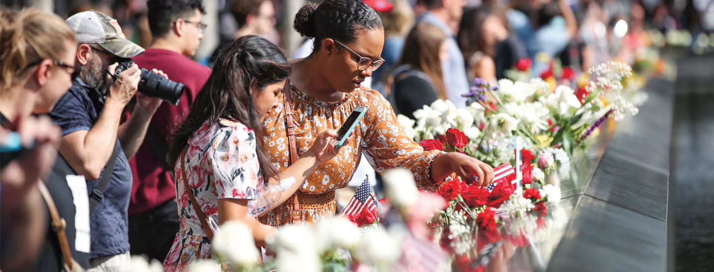 A crowd gathers at a memorial wall lined with flowers and small American flags